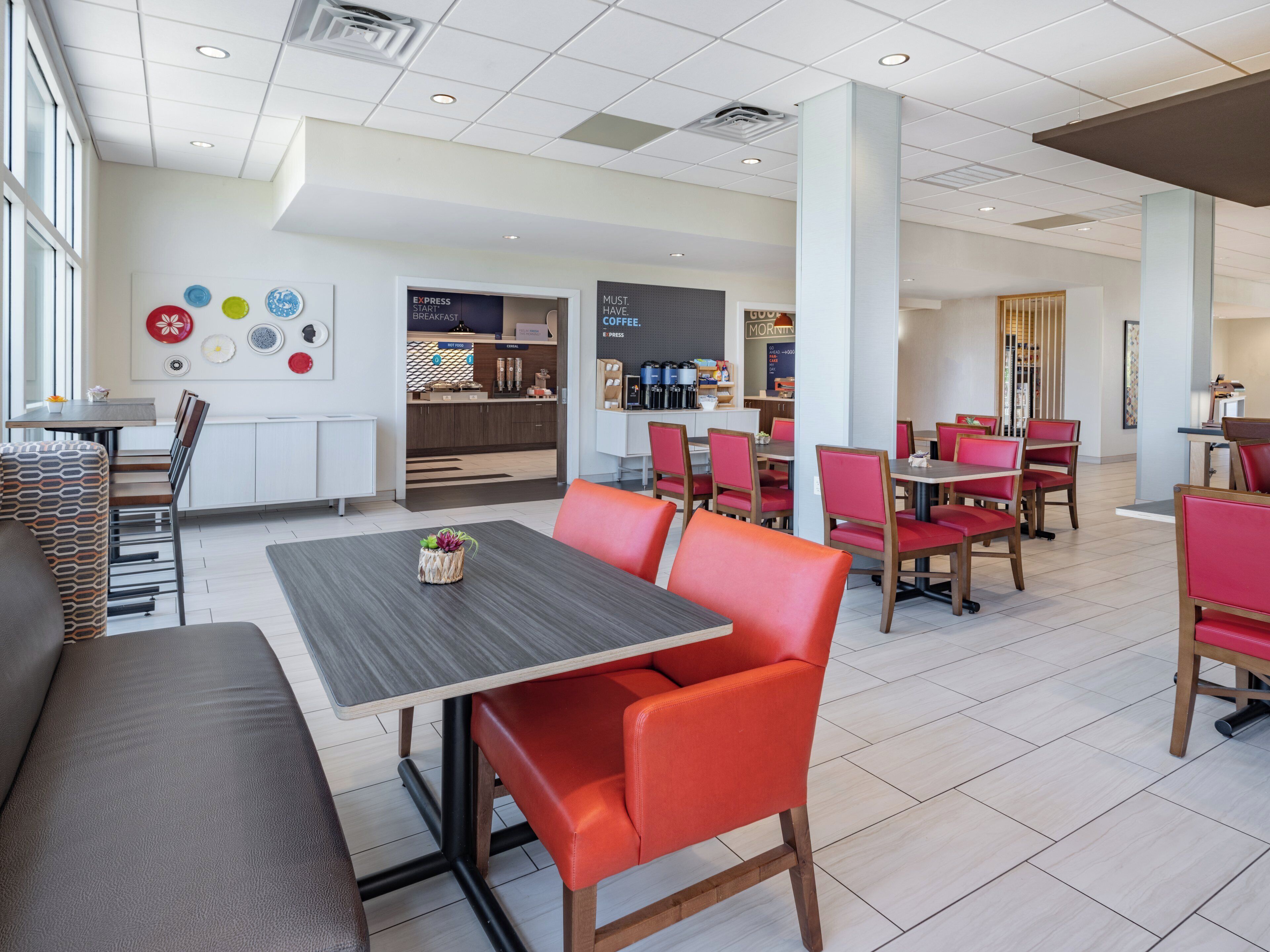 Restaurante moderno com área de refeições central, com cadeiras estofadas em vermelho e mesa de madeira, cercada por cozinha e balcão de café da manhã.