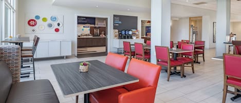 Restaurante moderno com área de refeições central, com cadeiras estofadas em vermelho e mesa de madeira, cercada por cozinha e balcão de café da manhã.