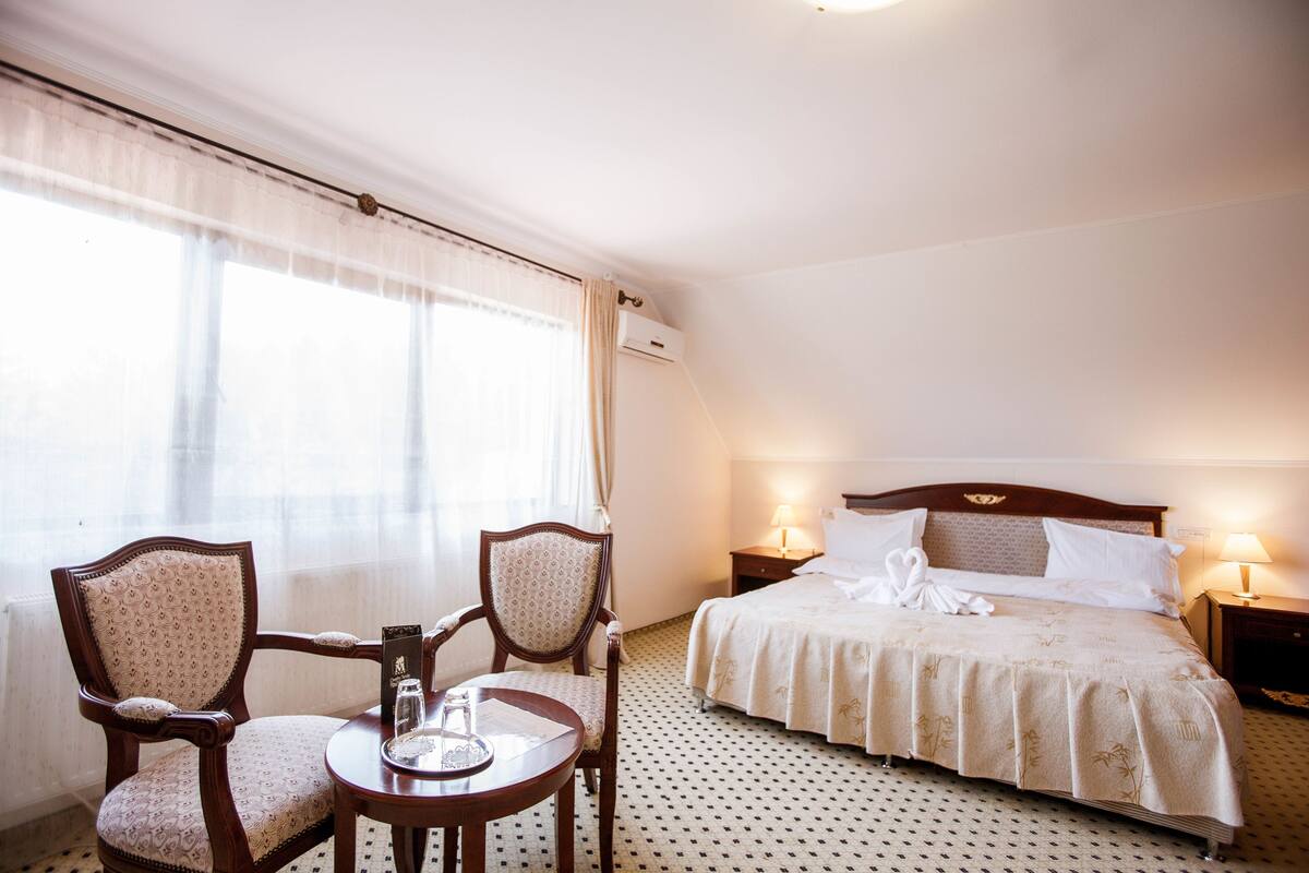 Hotellihuone, jossa on sänky, kaksi tuolia, pieni pöytä ja yöpöytä, jossa on lamput.