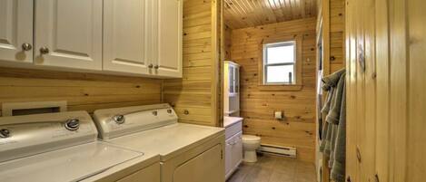 A compact, wooden cabin-style bathroom with a washing machine, a toilet, and a window.