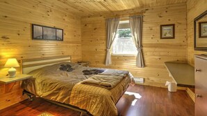 Et soveværelse i en træhytte med seng, et sengebord med lampe, et vindue med gardiner og en lille kommode.