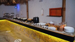 Montaje de buffet con diversos alimentos y recipientes, una pared de piedra y un techo de paja.