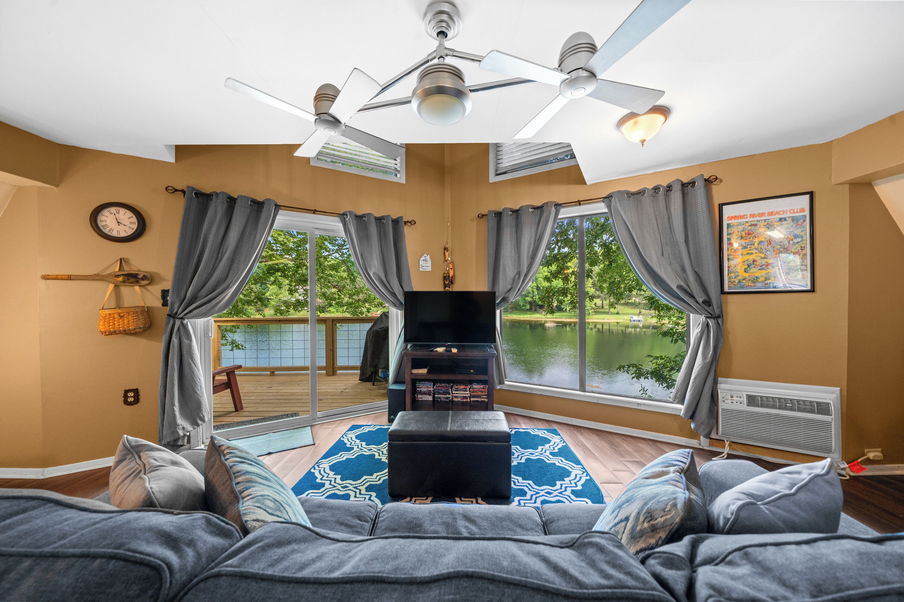 A room with a large window, a ceiling fan, a flat-screen TV, and a view of a lake.