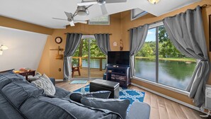 Un salón con sofá, televisor, ventilador de techo y ventanas amplias con vistas al agua.