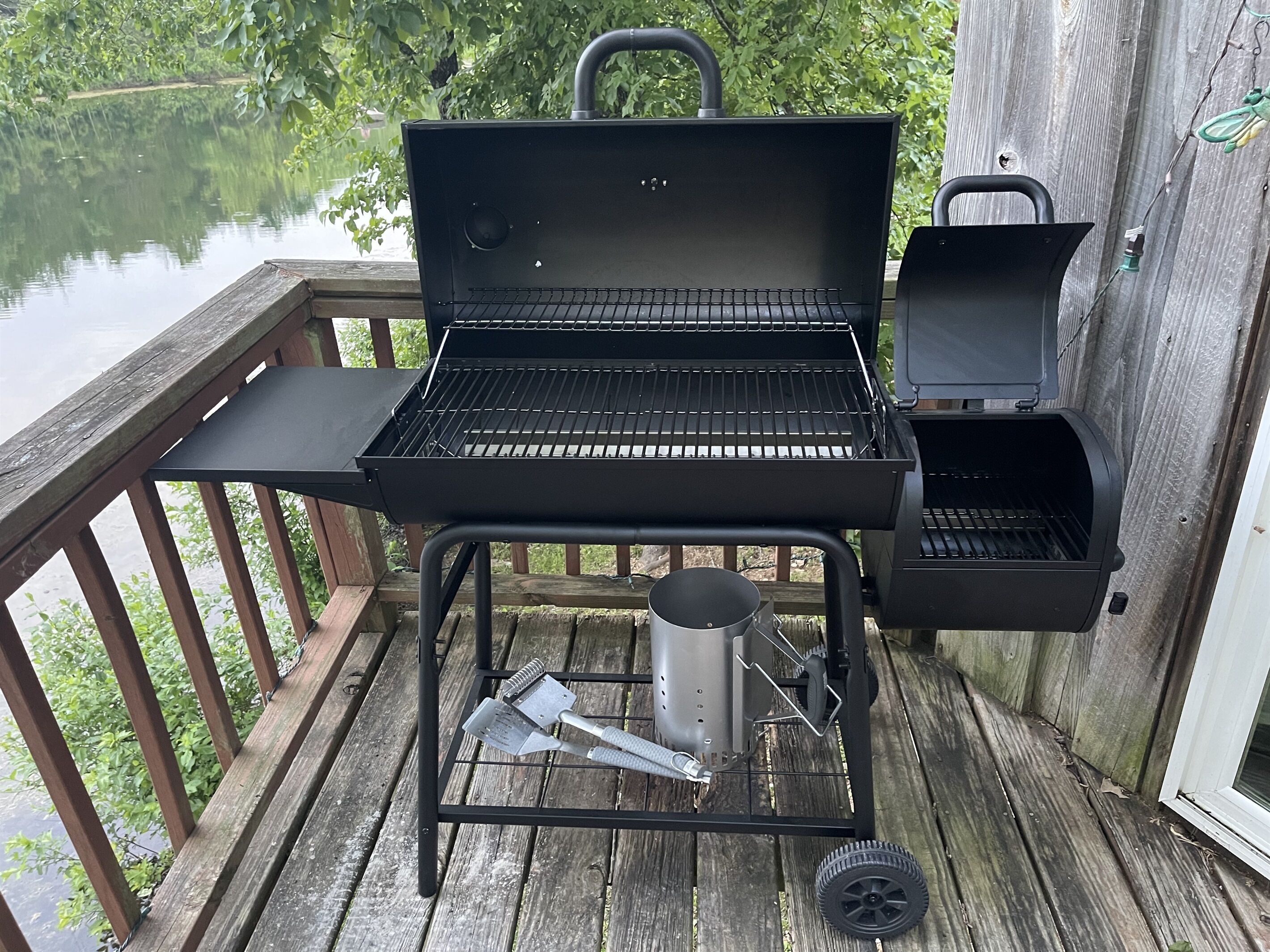 A portable grill with a side burner and a built-in smoker, placed on a wooden deck with a view of a lake.