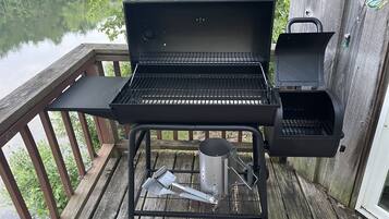 A portable grill with a side burner and a built-in smoker, placed on a wooden deck with a view of a lake.