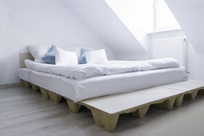 Une chambre de style loft moderne, avec un plafond en pente, un lit recouvert de linge de maison blanc et une plateforme en béton.