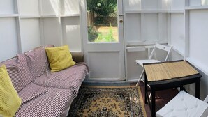 A small, enclosed room with a white door, a striped sofa with yellow cushions, a small table with a woven mat, and a patterned rug. - London Walthamstow Home (London)
