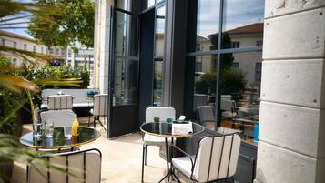 Un patio al aire libre con mesas y sillas redondas, ventanas amplias y vistas de la ciudad.