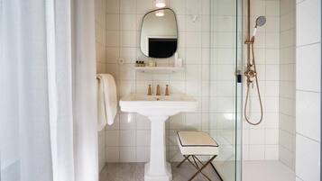 Un baño con lavamanos pedestal, espejo, toallero, cabina de ducha de cristal y un taburete pequeño.