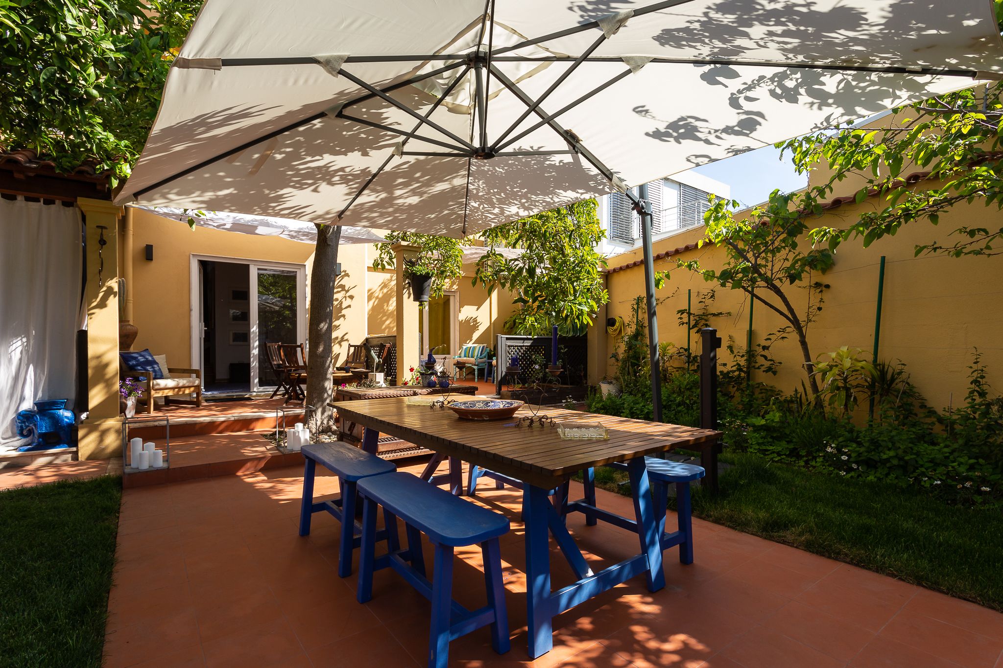 Un patio al aire libre con una mesa de madera y bancos azules, rodeado por un muro amarillo y follaje.