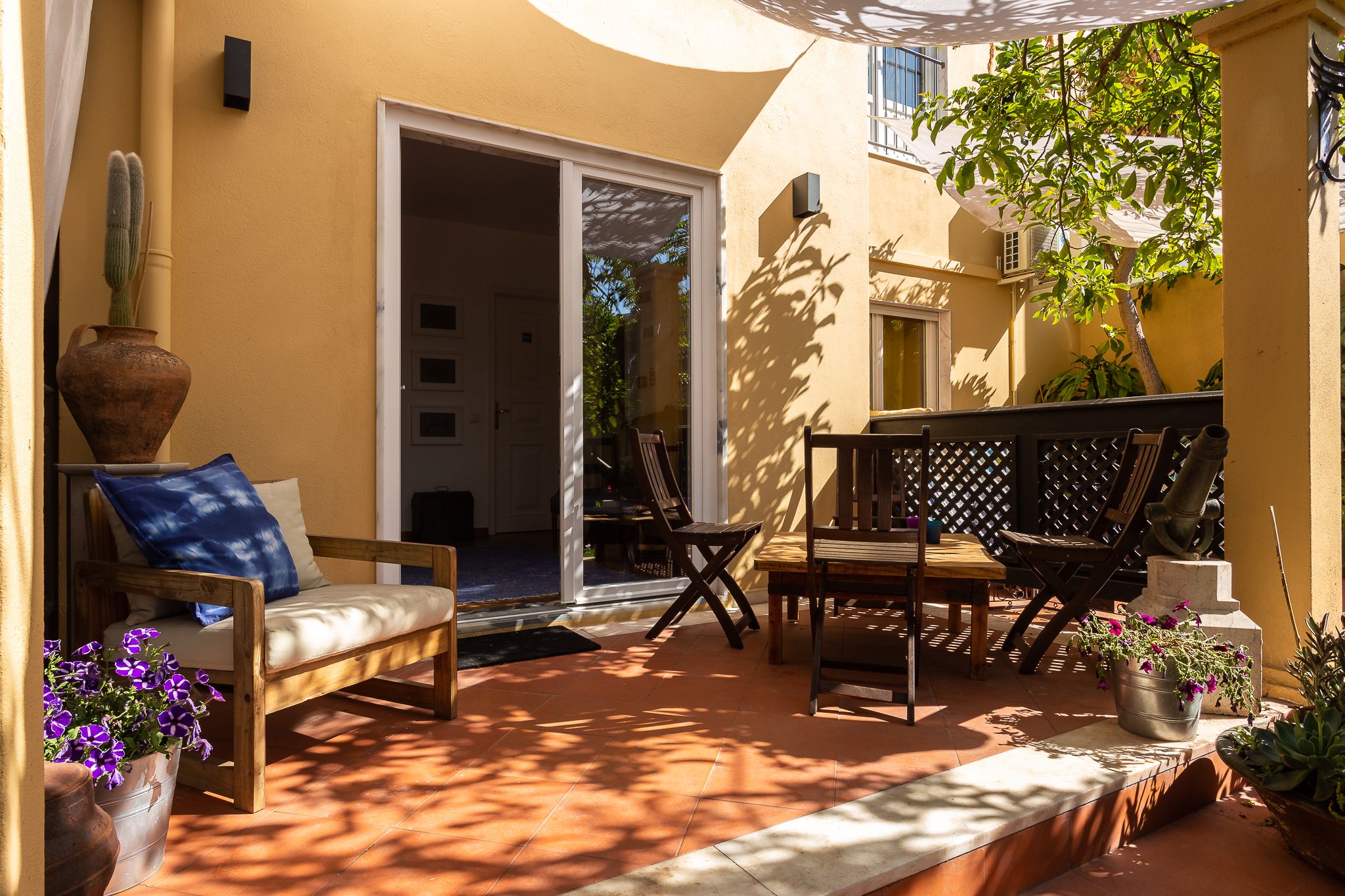 A sunlit patio with a wooden bench, a table, and chairs, surrounded by potted plants and a cactus.