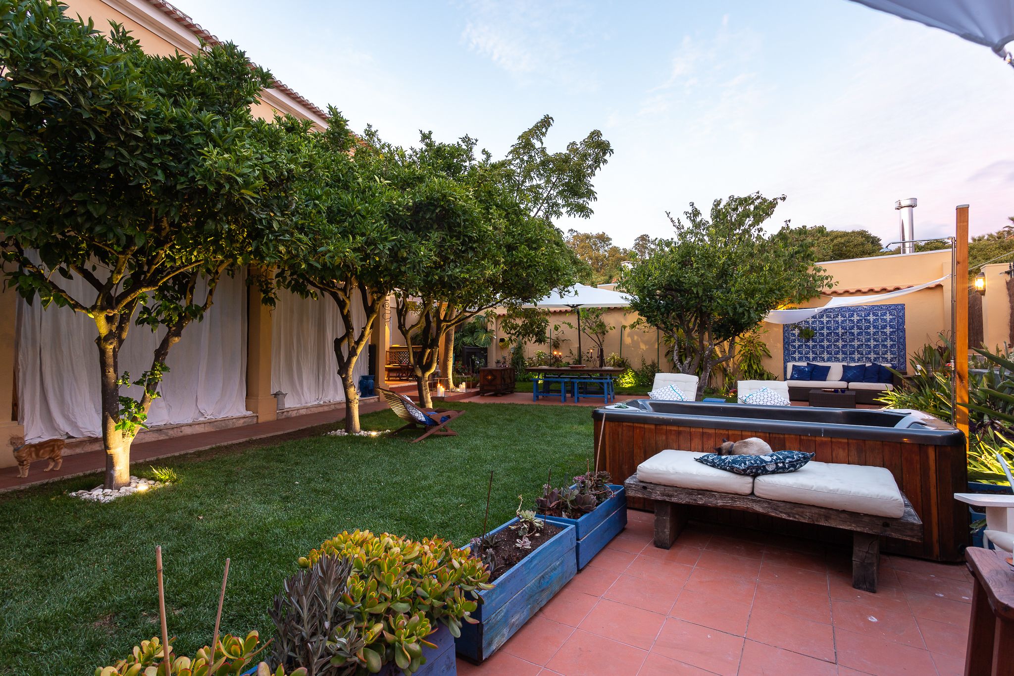 A patio area with a wooden bench, a small table, and a hot tub. There are potted plants and a tiled floor.