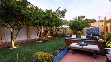 A patio area with a wooden bench, a small table, and a hot tub. There are potted plants and a tiled floor.