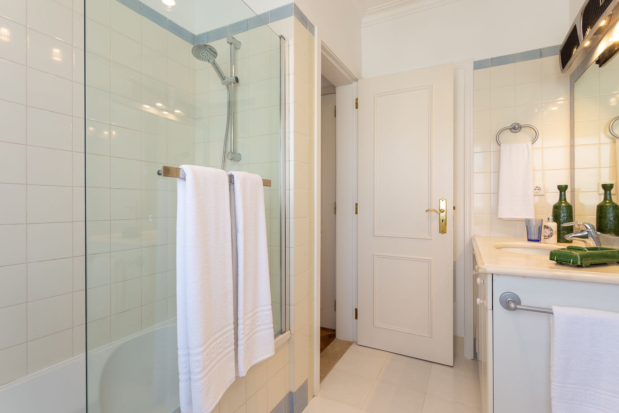 Une salle de bain moderne avec une douche à parois de verre, des carreaux blancs, un lavabo avec un miroir et une porte donnant sur une autre pièce.