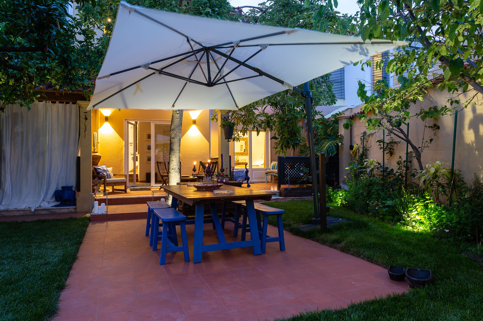 Un patio exterior con mesa y bancos, un paraguas grande y un interior luminoso visible a través de una puerta abierta.