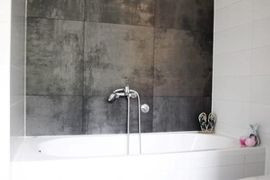 Een moderne badkamer met een groot bad, een douchekop en een paar slippers op de rand.