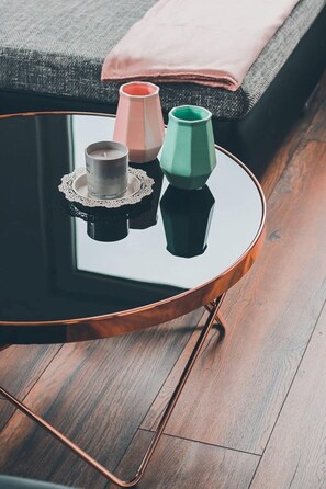 Een ronde glazen salontafel met een koperen voet, een klein metalen bekertje en twee keramieken vazen op een houten vloer.