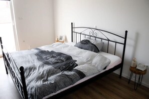 Een bed met een metalen frame, een nachtkastje met een lamp en een klein houten kruk.