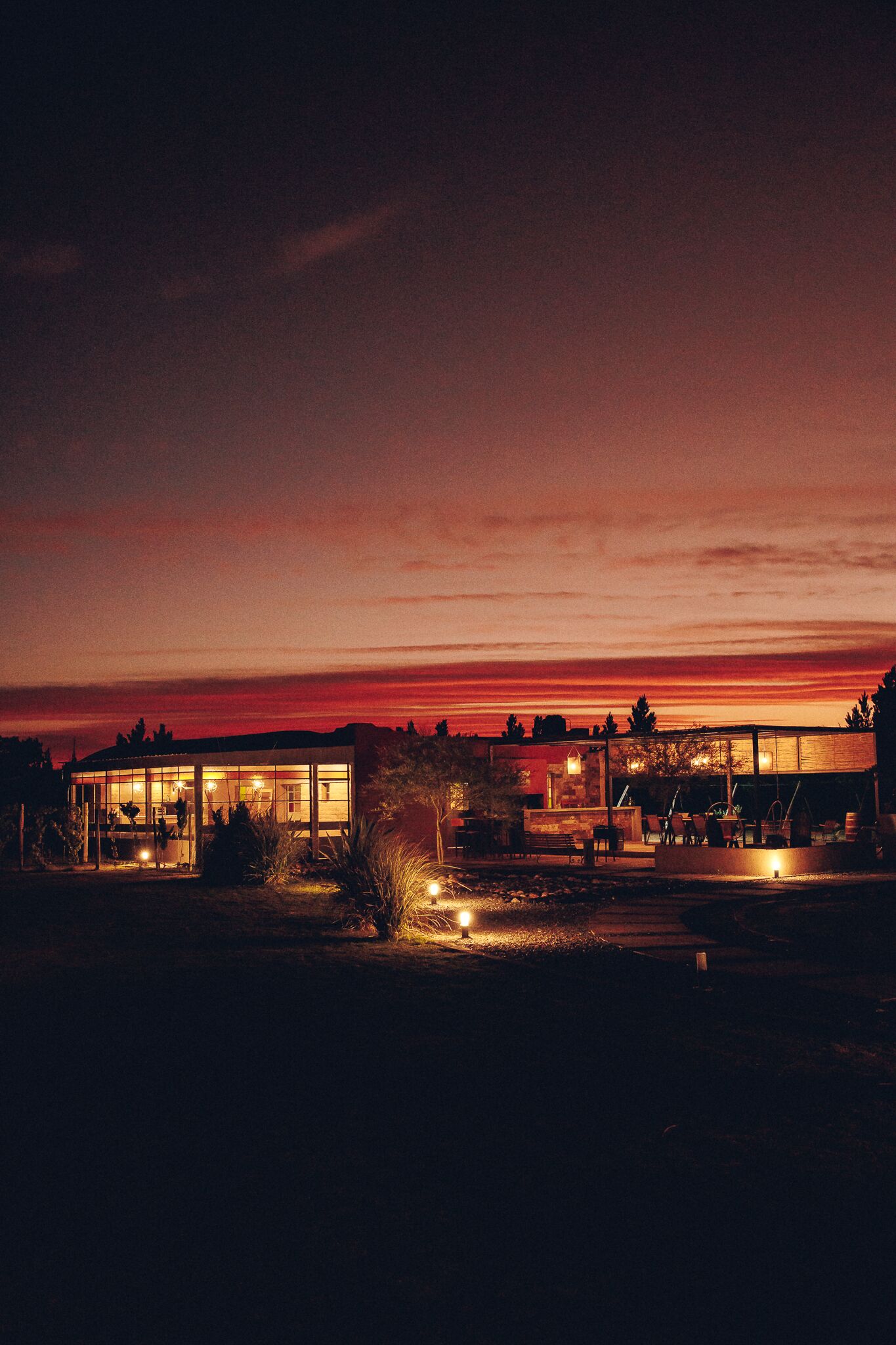 A modern house with large glass windows, a well-lit outdoor area, and a sunset sky.