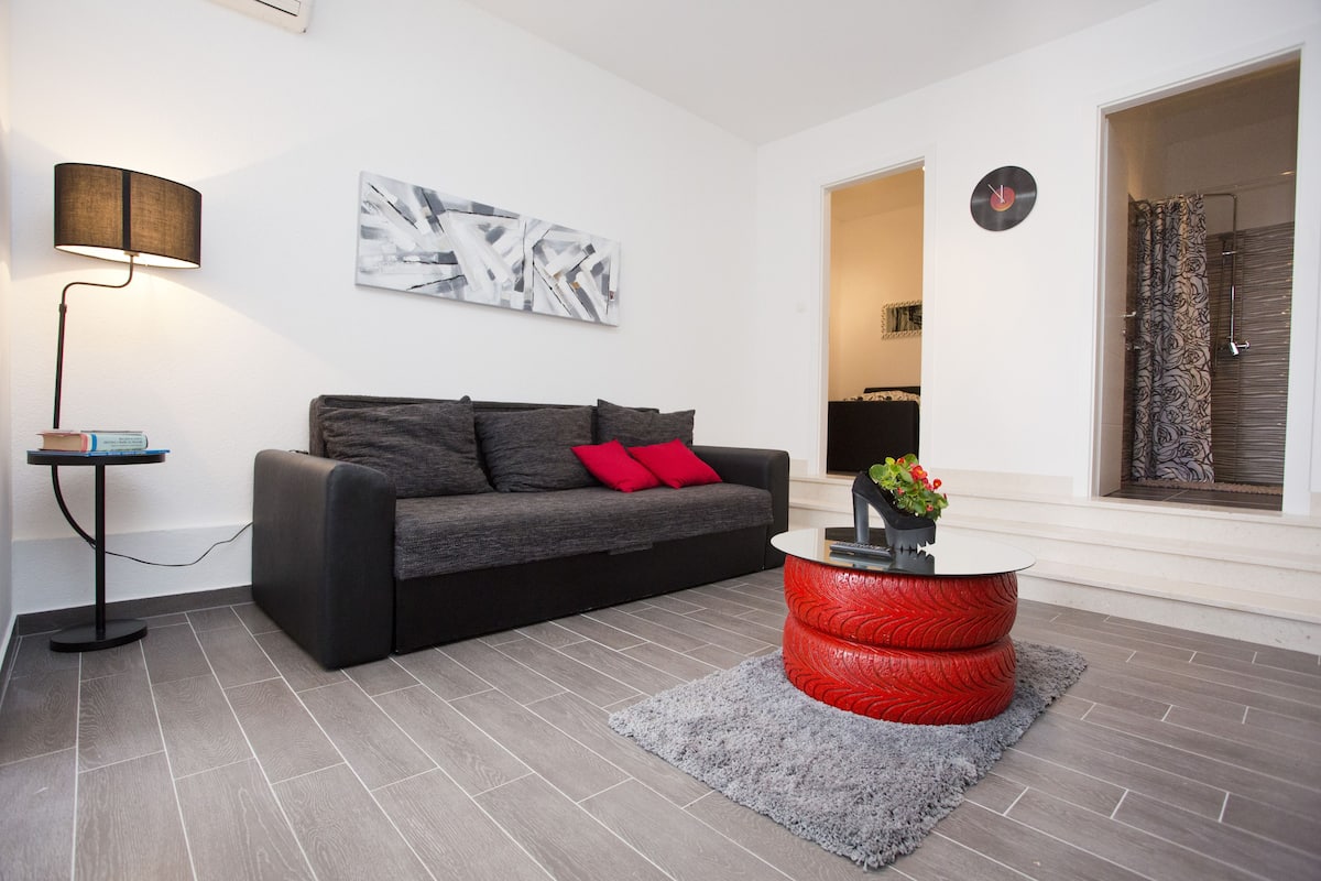 A modern living room with a grey sofa, red cushions, a round red coffee table, a grey rug, a floor lamp, and a wall clock.