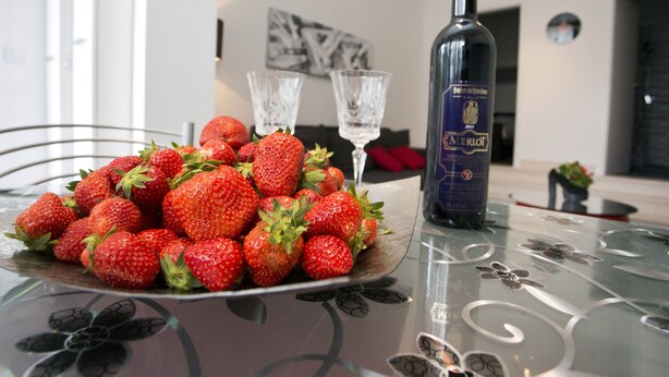 Una mesa de comedor con un plato de fresas, dos copas de vino y una botella de vino Merlot.