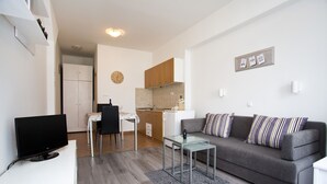 Una sala de estar moderna con un sofá gris, una mesa de centro de vidrio y un televisor de pantalla plana. Hay una zona de cocina con armarios de madera y una encimera blanca.