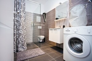 A modern bathroom with a glass shower enclosure, a toilet, a washing machine, and a vanity with a mirror. - Apartments Silver Tree (Split)
