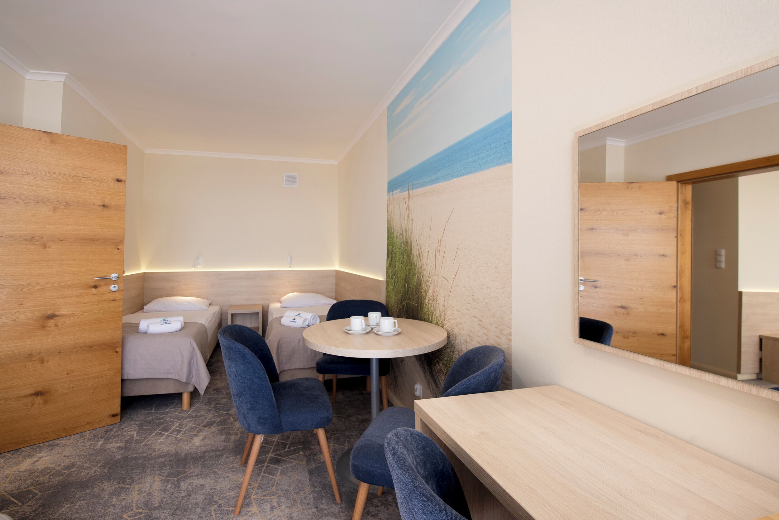 Una habitación de hotel con cama, dos sillas, una mesita con tazas, un espejo y una puerta de madera.