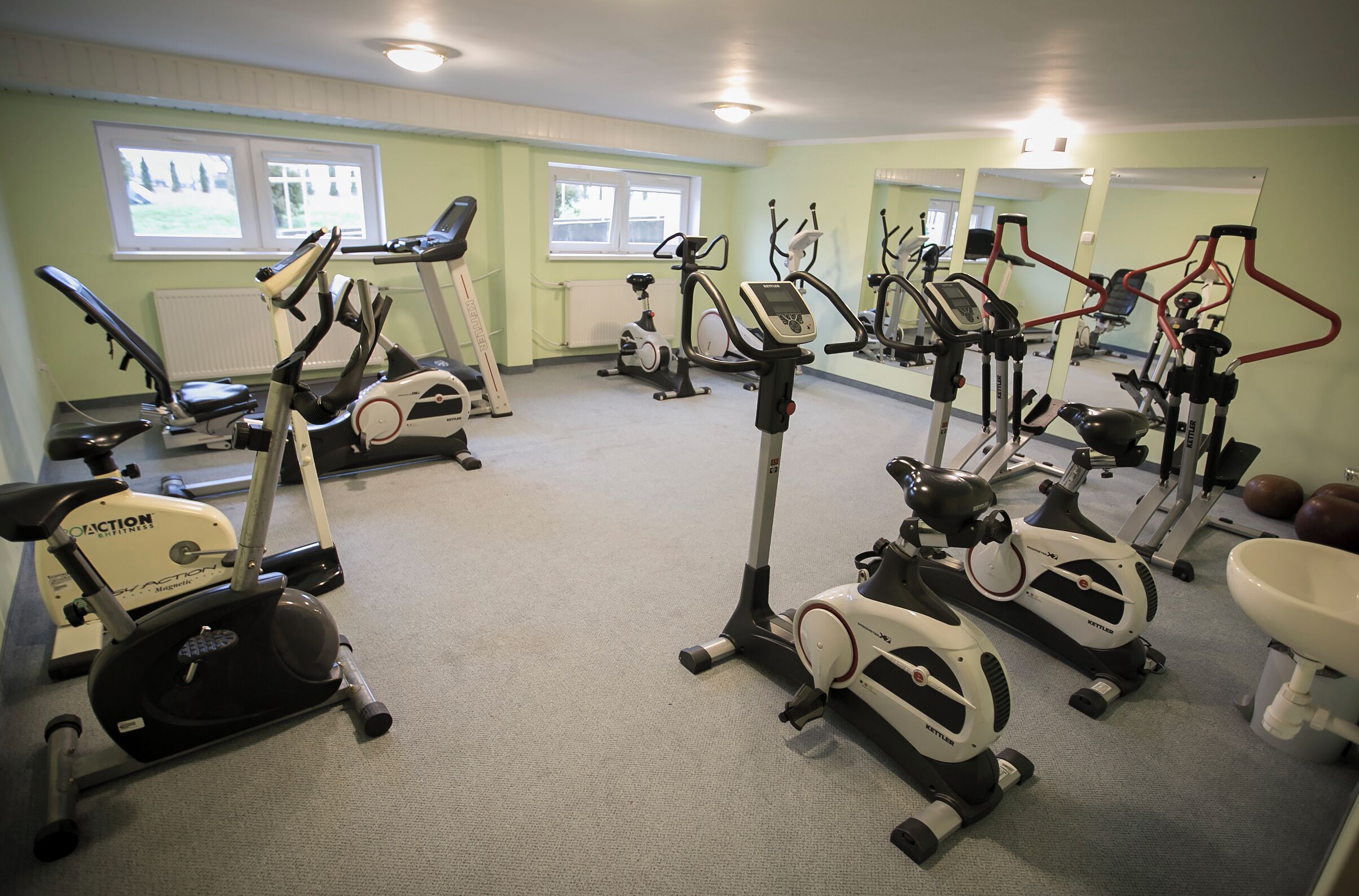 Un gimnasio con varias máquinas de ejercicio, un espejo grande y una ventana con vista a árboles.