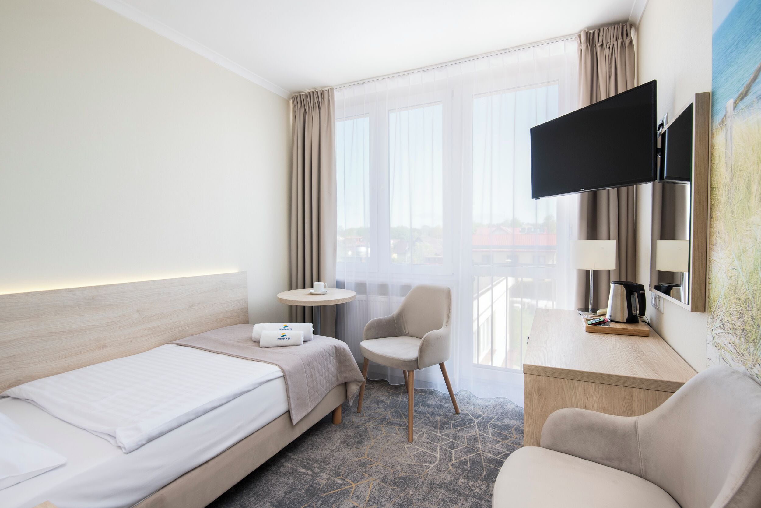A hotel room with a bed, a desk with a flat-screen TV, a chair, a small round table, and a window with curtains.