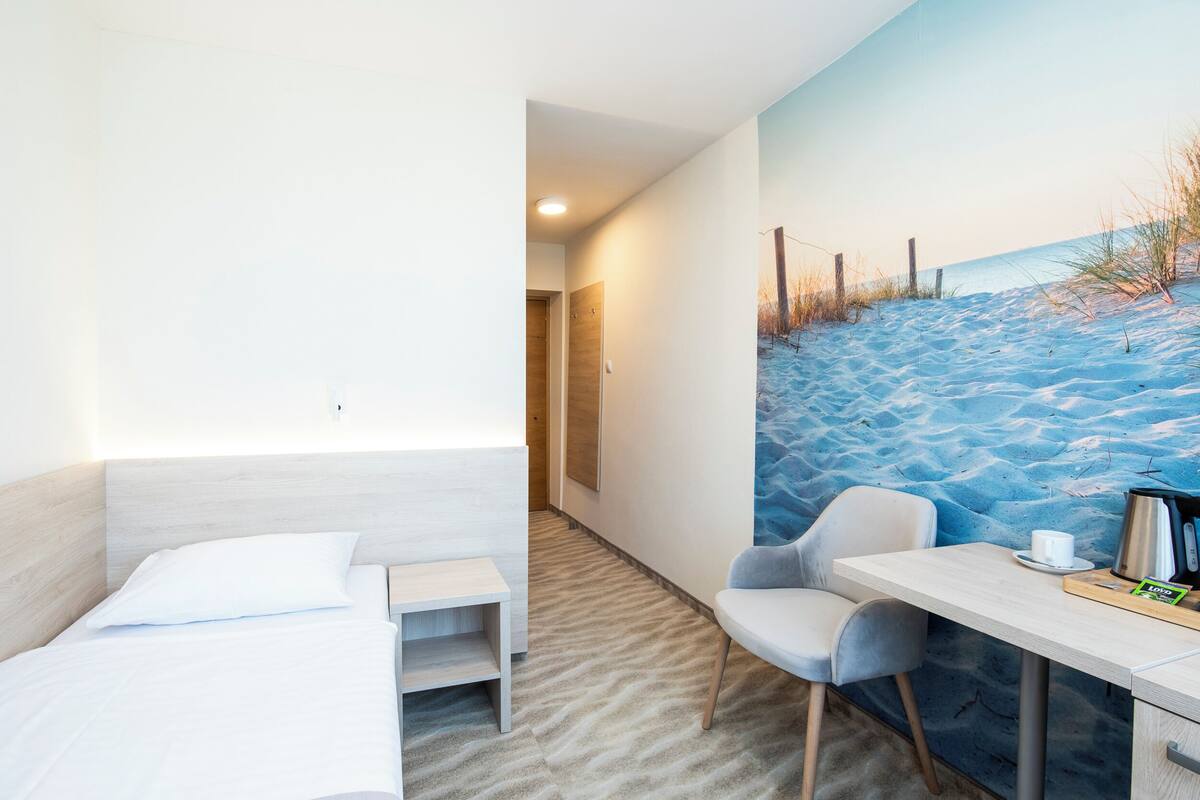 A room with a bed, a small table, a chair, and a painting of a beach scene.