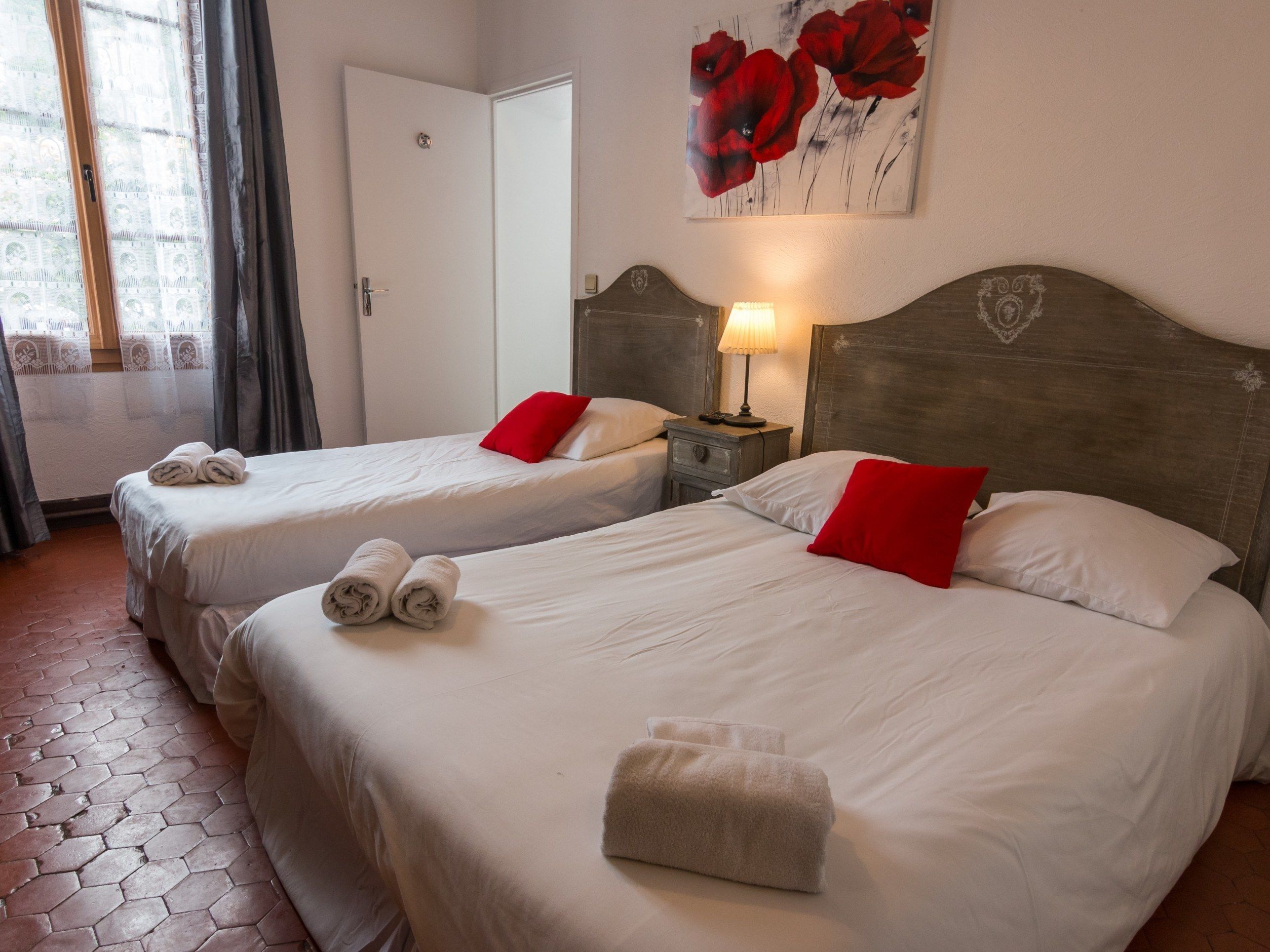 Camera d'albergo con due letti, ognuno con lenzuola bianche e cuscini rossi, un comodino con una lampada e una porta a sinistra.