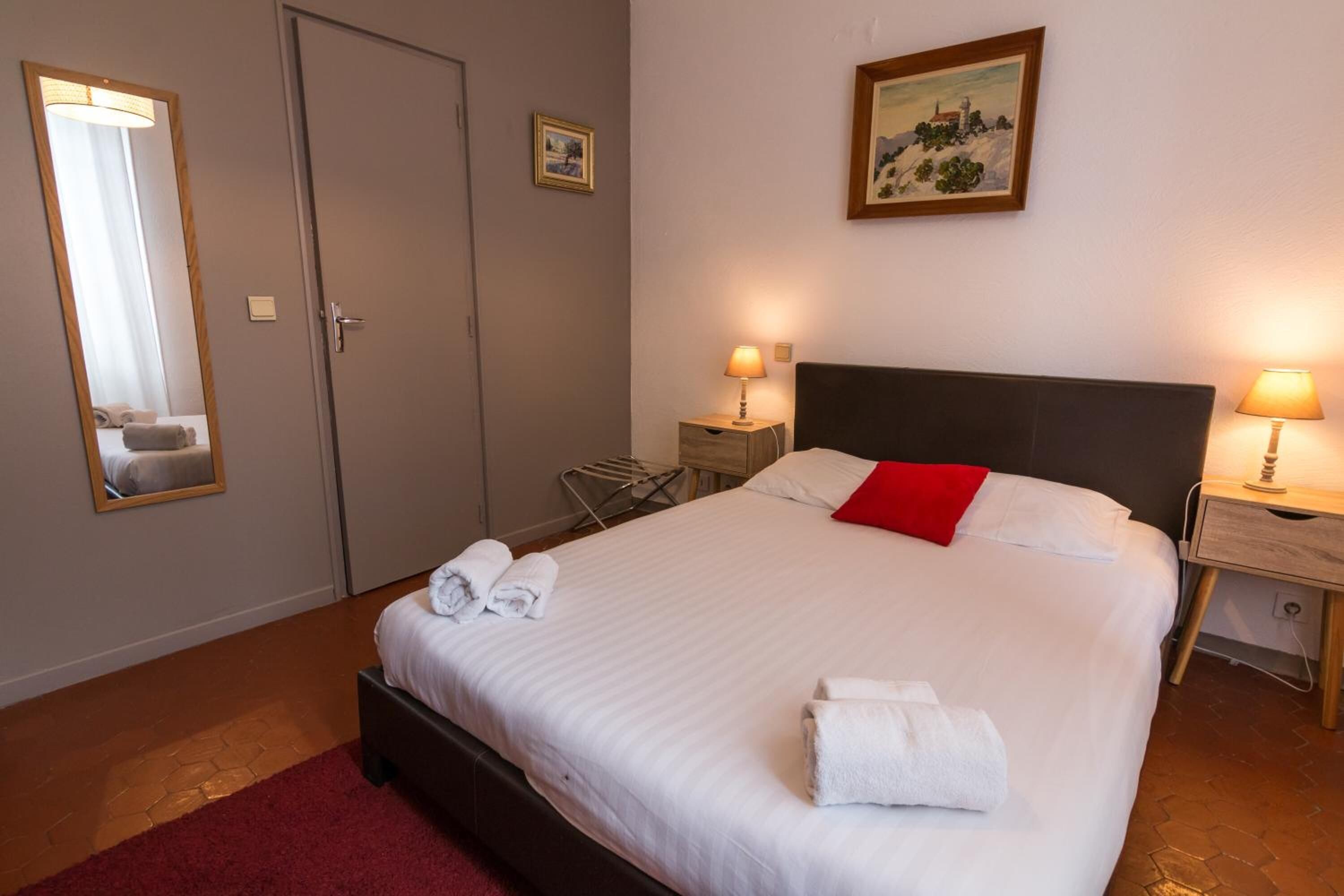 Una cama bien hecha con sábanas blancas y una almohada roja, dos lámparas de noche, un cuadro en la pared y una puerta con un perchero al lado.