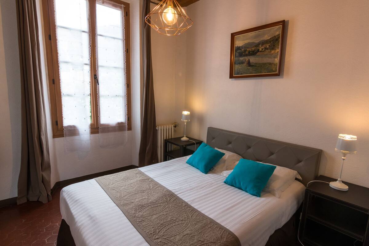 Una cama bien hecha con una colcha blanca y color beige, dos almohadas azules y un cabecero gris. Hay un cuadro en la pared sobre la cama y una lámpara en la mesita de noche a su lado.