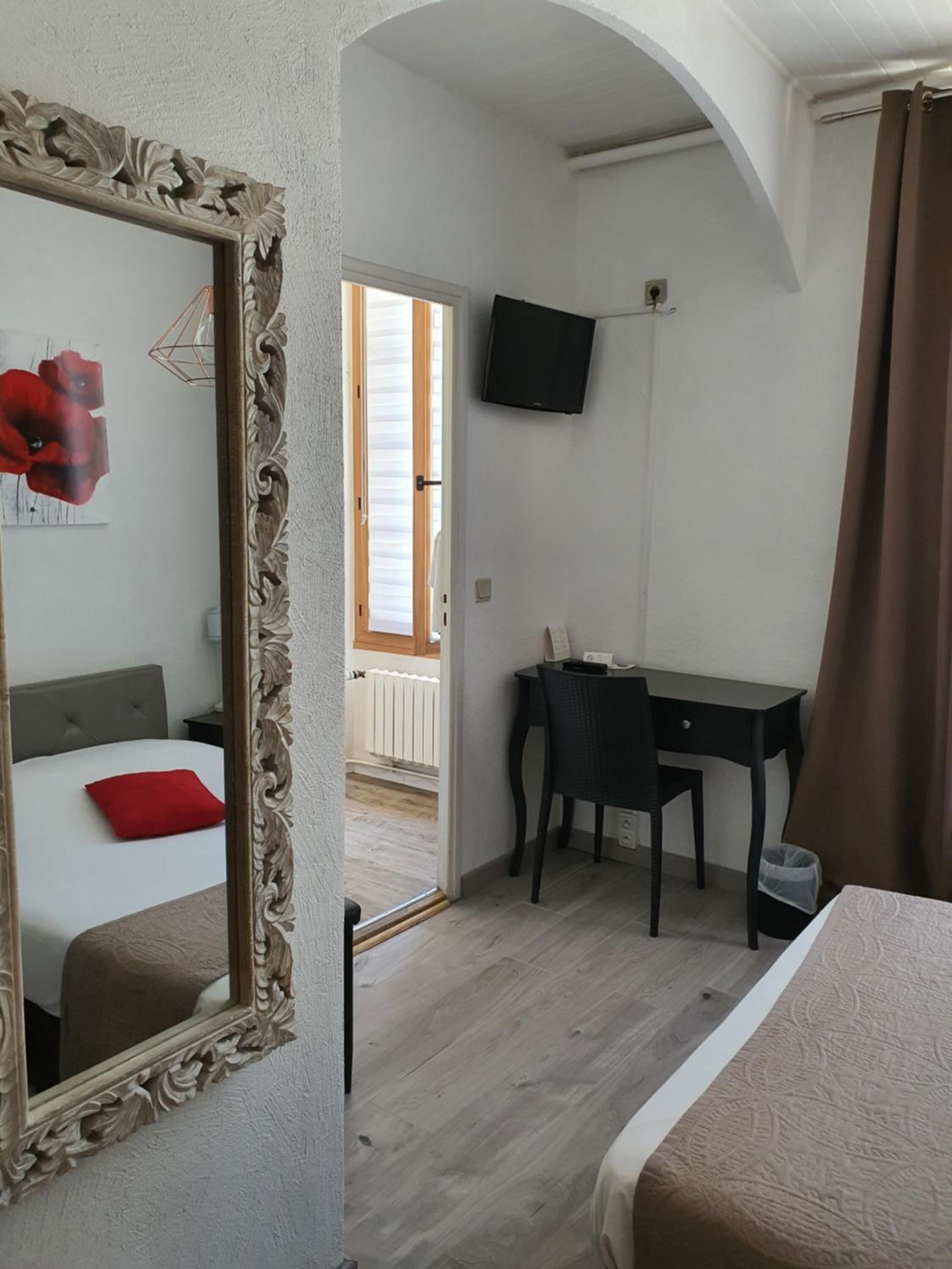 A bedroom with a large mirror, a bed with a red pillow, a desk, and a television on the wall.