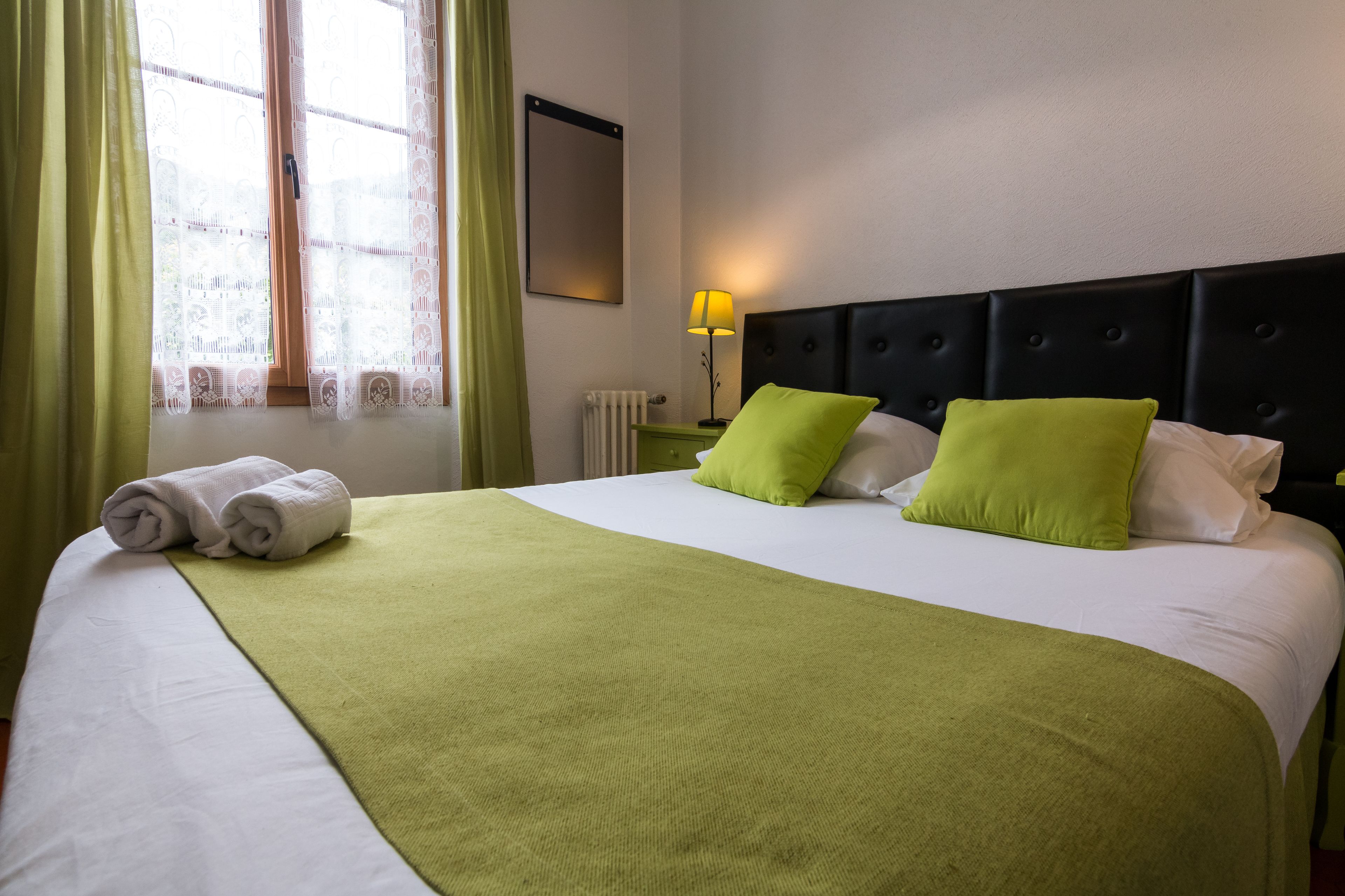 A neatly made bed with a green bedspread and two pillows, a nightstand with a lamp, and a window with green curtains.