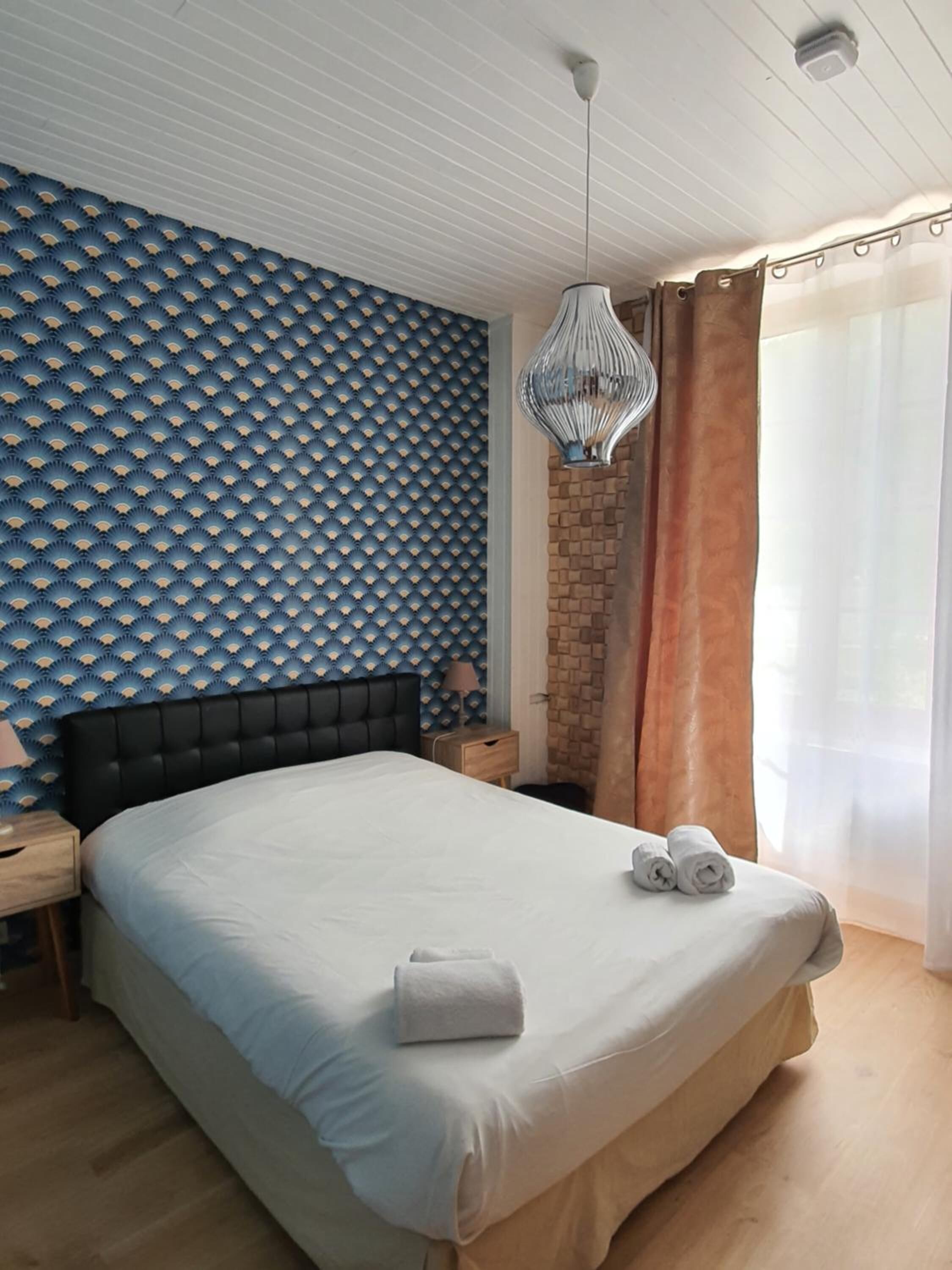 A bedroom with a patterned wall, a bed with white linens, a nightstand, a lamp, and a hanging light fixture.