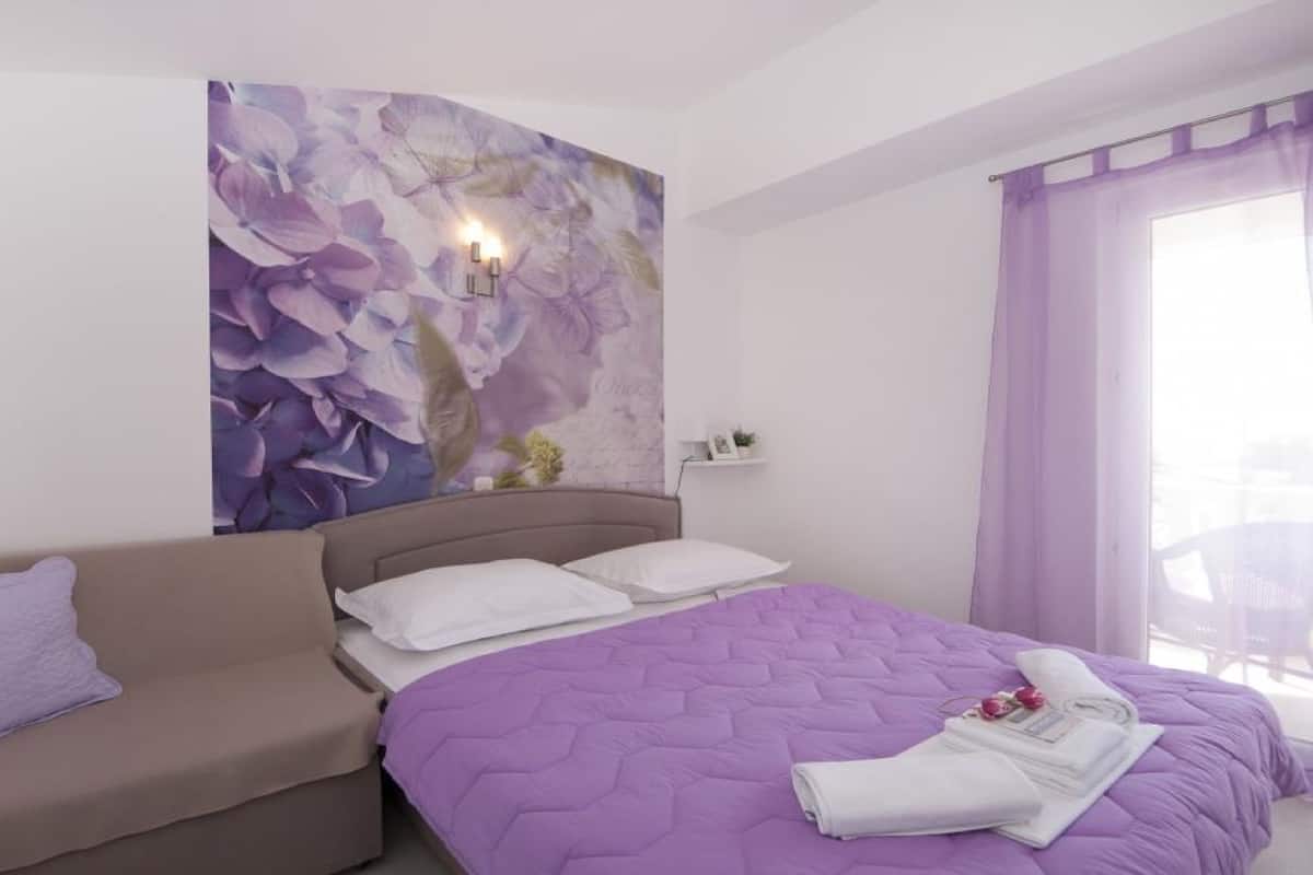 A bedroom with a bed, a sofa, a wall with a floral mural, and a window with purple curtains.