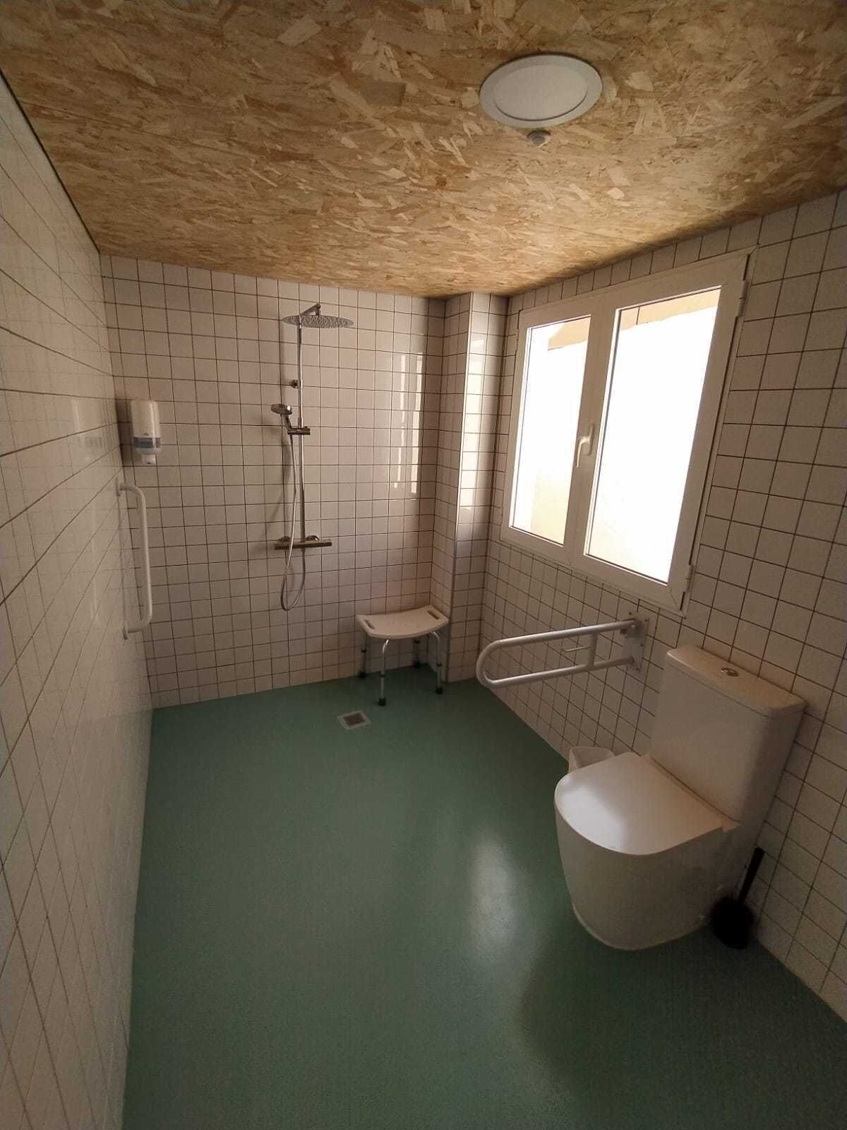 A small, tiled bathroom with a green floor, a shower, a toilet, and a window.