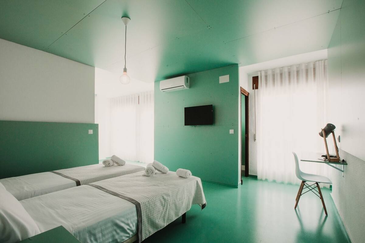 A hotel room with two beds, a desk, a chair, a TV, and an air conditioning unit.
