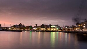 Un puerto deportivo al anochecer con barcos amarrados, edificios iluminados y una superficie acuática tranquila que refleja las luces.
