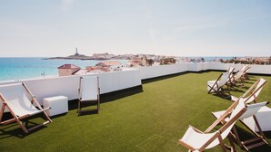 Una terraza en la azotea con césped artificial, sillones blancos y vistas al mar y al paisaje urbano.