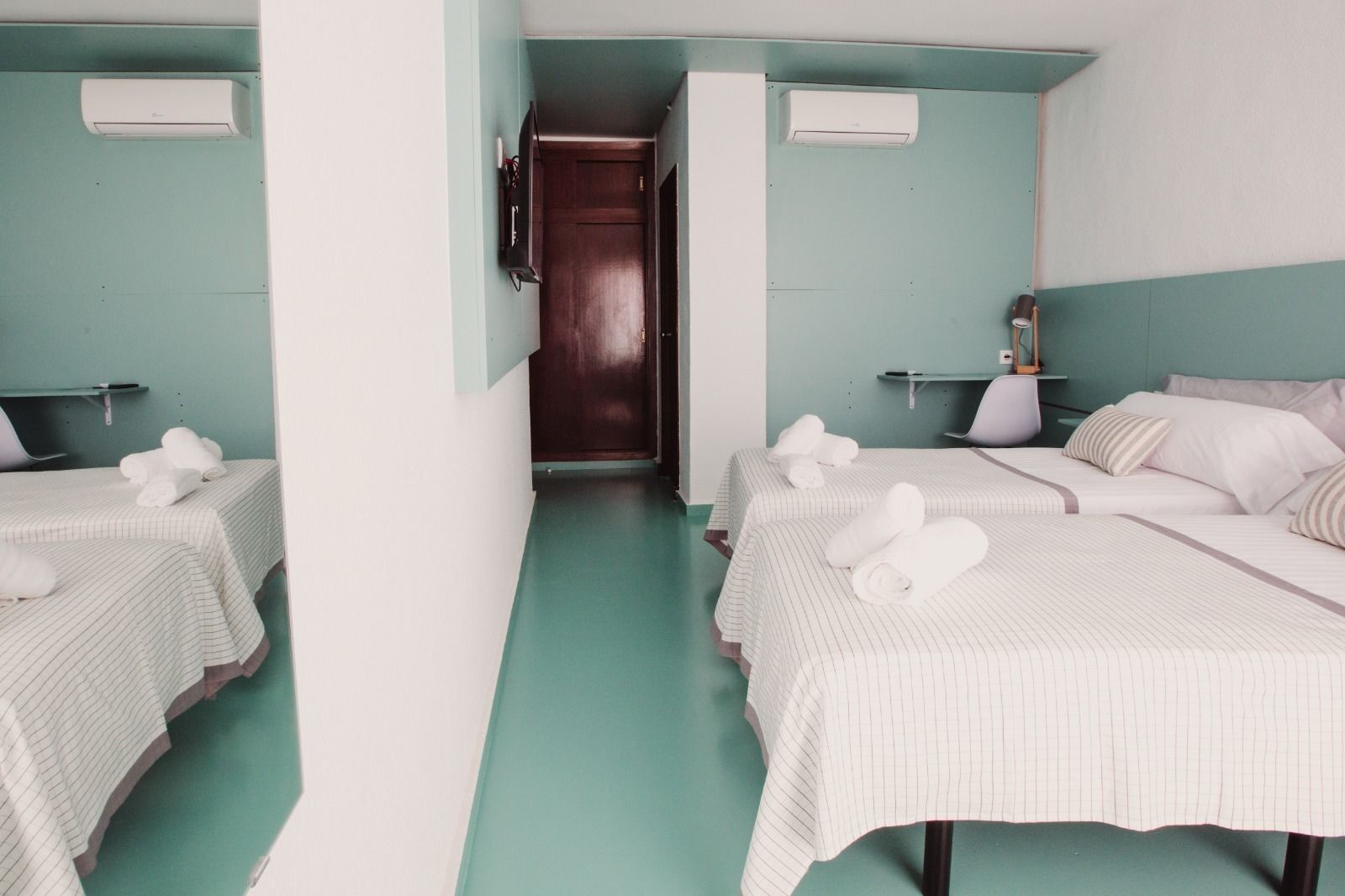 A hotel room with two beds, each with a white striped bedspread and rolled towels. There are air conditioning units on the walls, a wooden door, and a small table with a lamp.