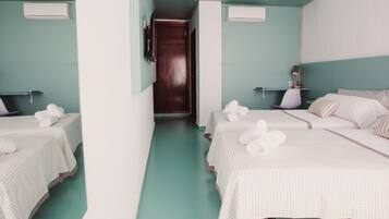 A hotel room with two beds, each with a white striped bedspread and rolled towels. There are air conditioning units on the walls, a wooden door, and a small table with a lamp.