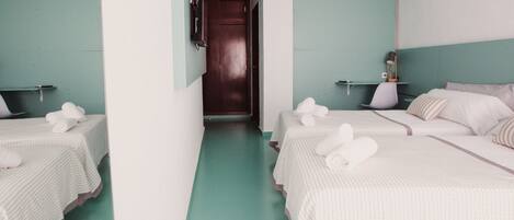 A hotel room with two beds, each with a white striped bedspread and rolled towels. There are air conditioning units on the walls, a wooden door, and a small table with a lamp.