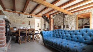 Una habitación rústica con vigas de madera a la vista, paredes de piedra, un sofá azul, una mesa de comedor con sillas y una chimenea.