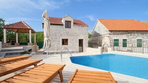 Un área junto a la piscina con tumbonas de madera, un edificio de piedra con tejado de tejas y una piscina azul cristalina.