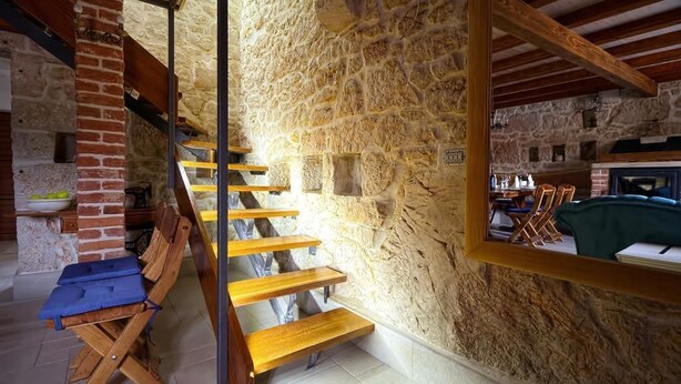 Un interior rústico de piedra con una escalera de madera, chimenea de ladrillo y comedor con mobiliario de madera.