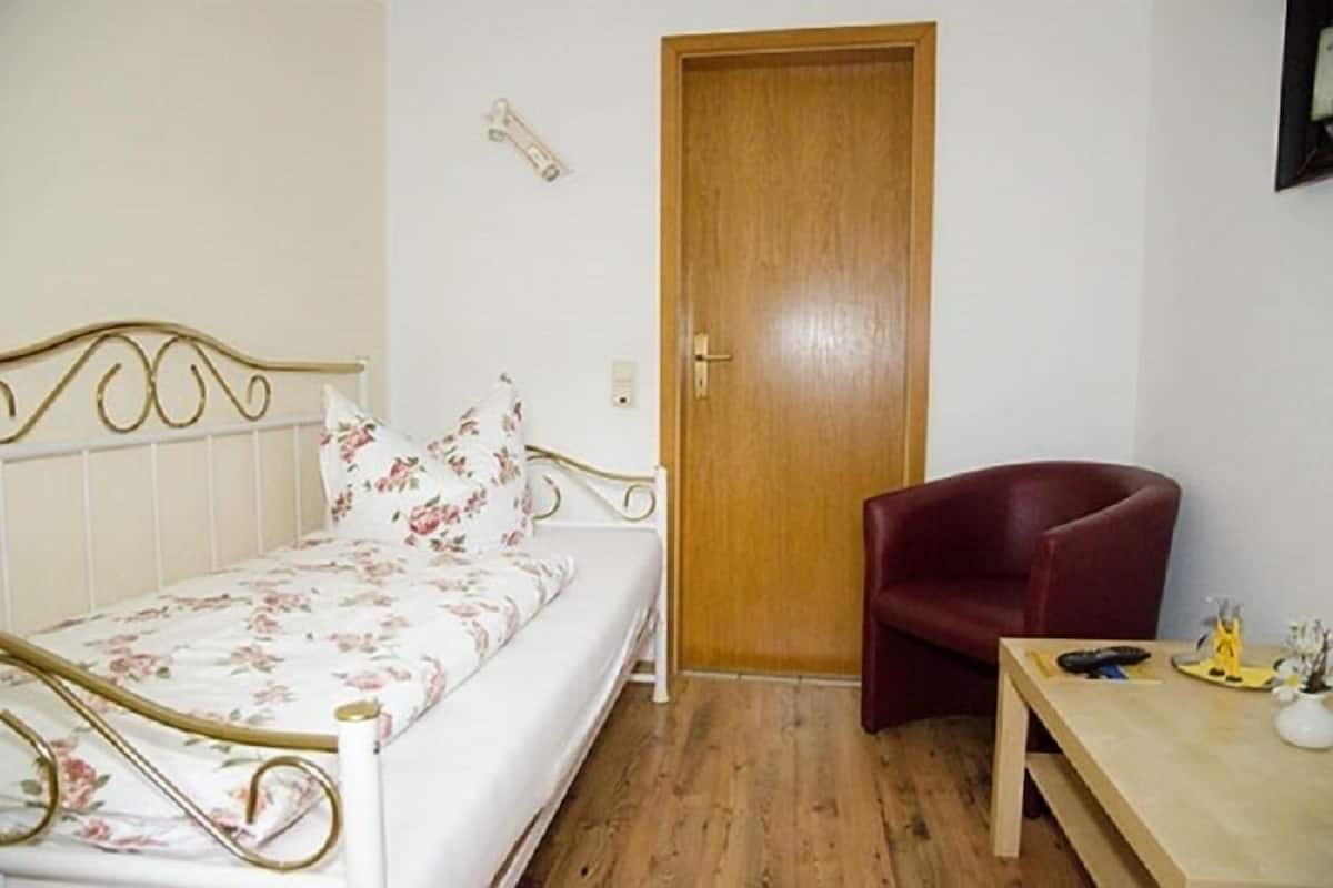 Un lit simple avec une couvre-lit à motifs floraux, une porte en bois, une chaise rouge et une petite table sur laquelle se trouvent un vase et une télécommande.